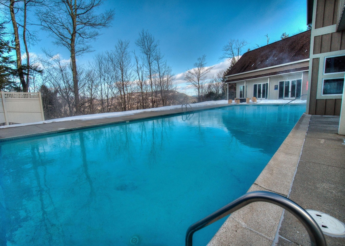 An outdoor heated pool is visible, surrounded by trees and offering views of the landscape. The water reflects the sky, and the area includes a concrete deck with a handrail leading into the pool. A building can be seen in the background.