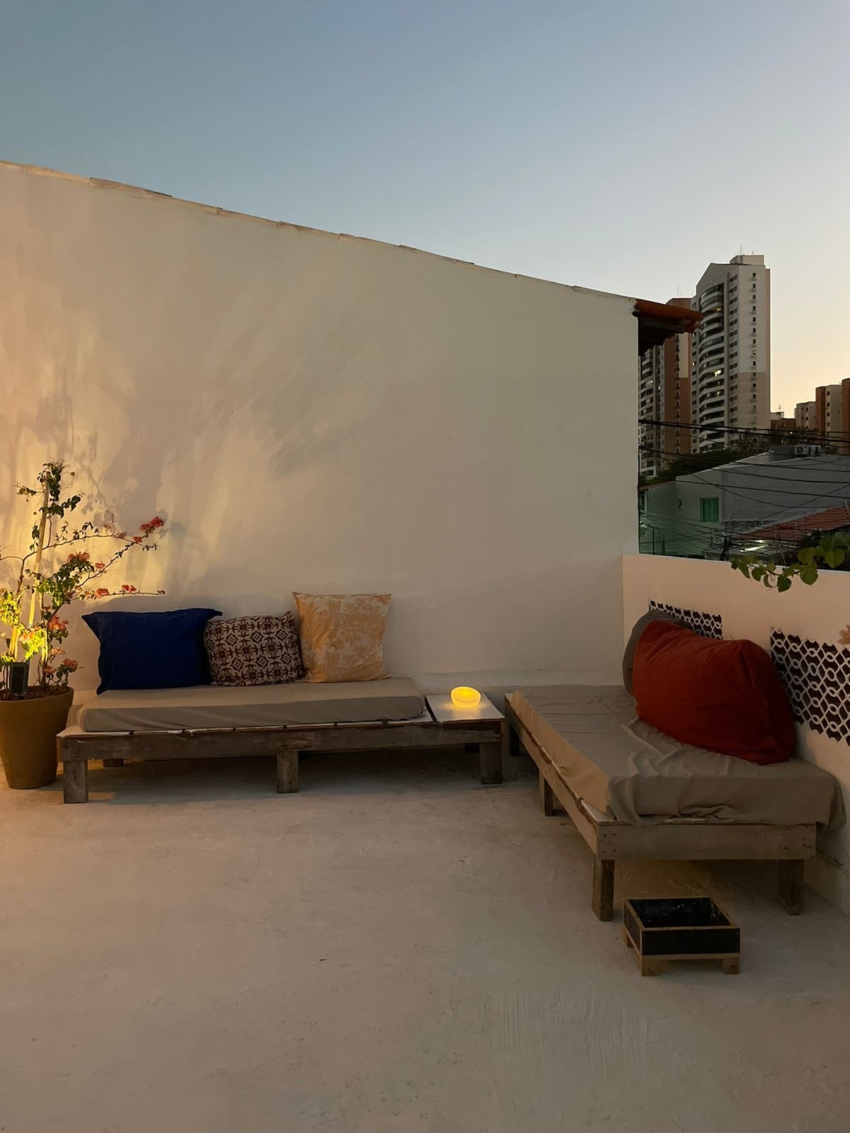 A peaceful terrace area is displayed, featuring two lounge seats with colorful cushions. A small potted plant adds greenery, and a soft glow from a lantern creates a relaxing atmosphere. The backdrop includes a glimpse of the city skyline, enhancing the outdoor experience.