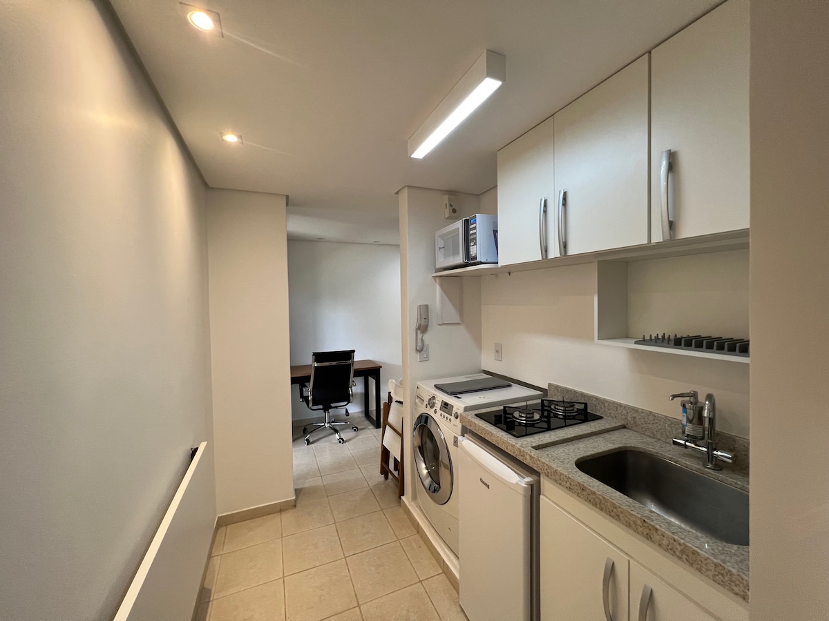 A bright and functional kitchen area is shown, featuring white cabinetry and modern appliances. A washing machine and kitchen equipment are visibly arranged. A separate workspace with a desk and chair can be seen in the background, highlighting the apartment's practicality.