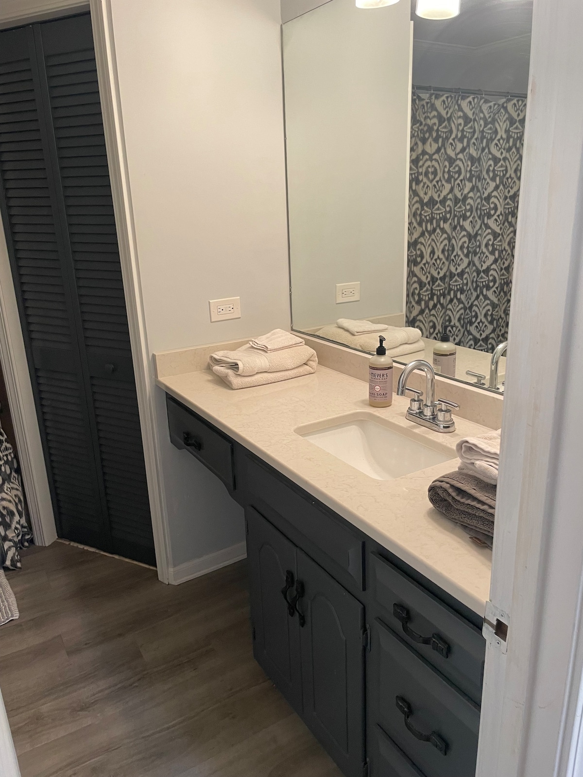 A serene bathroom is presented with a light-colored countertop and a sink. Towels are neatly arranged beside an elegant soap dispenser. A large mirror reflects the space, while textured wallpaper and dark cabinetry add a touch of contrast.