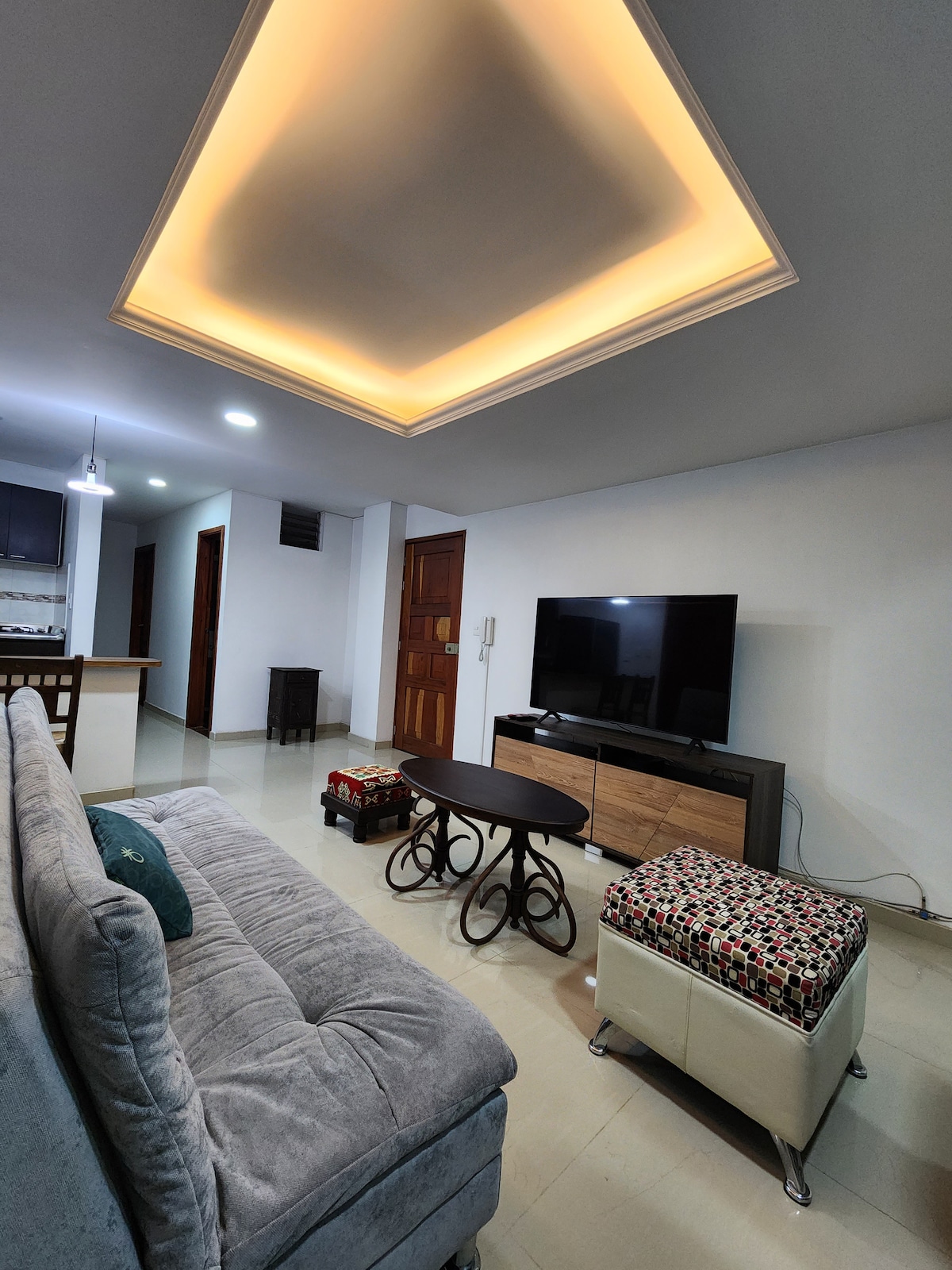 A modern living area displays a comfortable sofa and a decorative ottoman, complemented by a mounted television. Soft lighting from a unique ceiling design enhances the space, while a small table adds functionality. The open layout leads into the kitchen area, visible in the background.