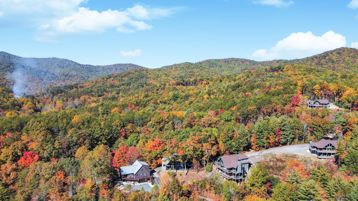 Aerial view captures the vibrant autumn foliage of the surrounding mountains, showcasing a palette of reds, oranges, and yellows. Several cabins are nestled among the trees, with winding roads visible beneath the expansive sky.