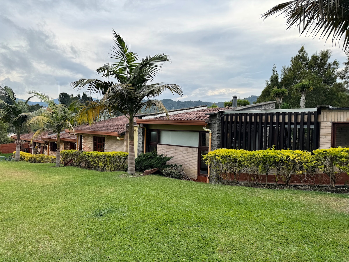 A serene outdoor landscape features a spacious lawn bordered by landscaped greenery and palm trees. Several adjoining buildings with terracotta roofs and a prominent wooden structure are visible, set against a backdrop of distant hills under an overcast sky.
