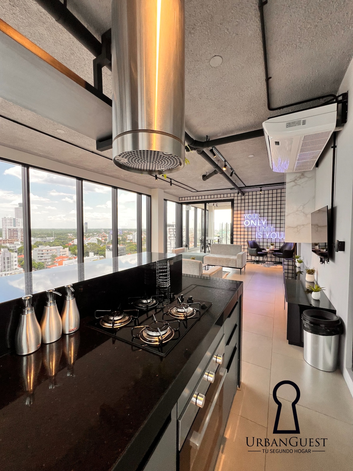 A modern kitchen is showcased, featuring sleek black countertops and stainless steel appliances. A five-burner gas stove is positioned under a stainless steel hood. Large windows provide a panoramic view of the urban landscape, flooding the space with natural light.