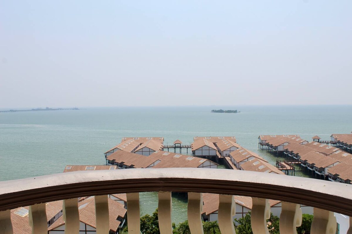 An expansive seaview stretches across the horizon, showcasing calm turquoise waters and wooden structures along the shoreline. A private balcony railing frames the view, enhancing the peaceful coastal ambiance.