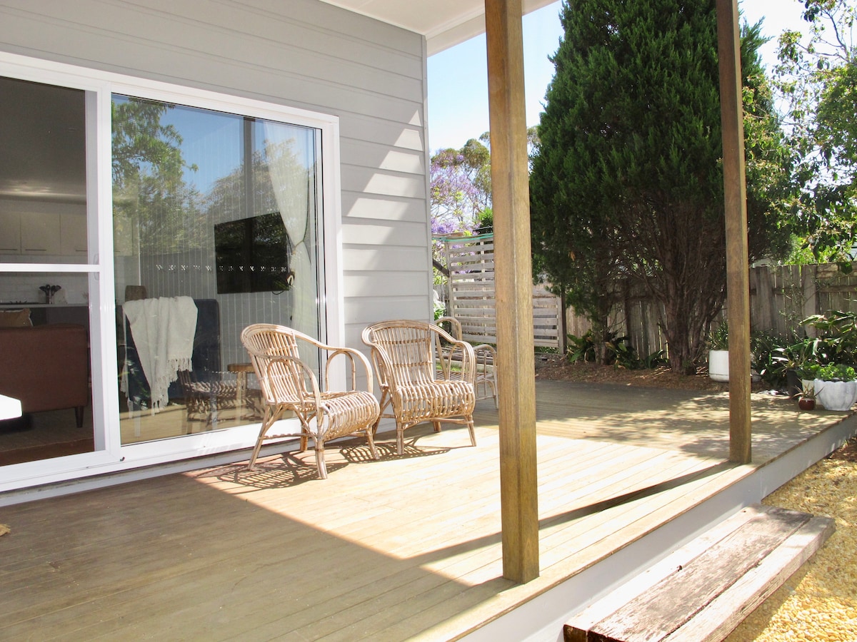 An outdoor deck area is seen with natural wood flooring, featuring two comfortable rattan chairs positioned next to a wall of glass. Lush greenery borders the space, providing a tranquil setting. Soft sunlight casts gentle shadows, enhancing the relaxed atmosphere.
