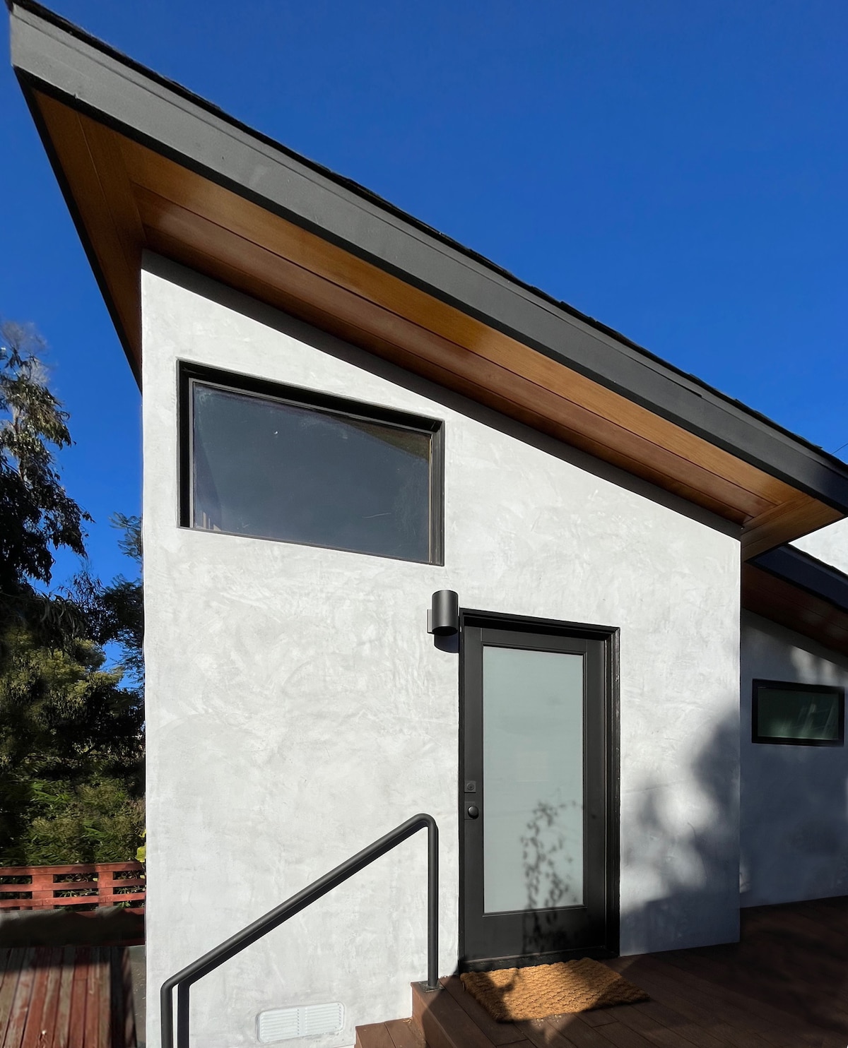 The exterior of the studio apartment features a modern design with a smooth, light-colored stucco finish. Large windows are positioned to optimize natural light, and a stylish front door is framed by an overhanging roof. A staircase leads to the welcoming entrance.