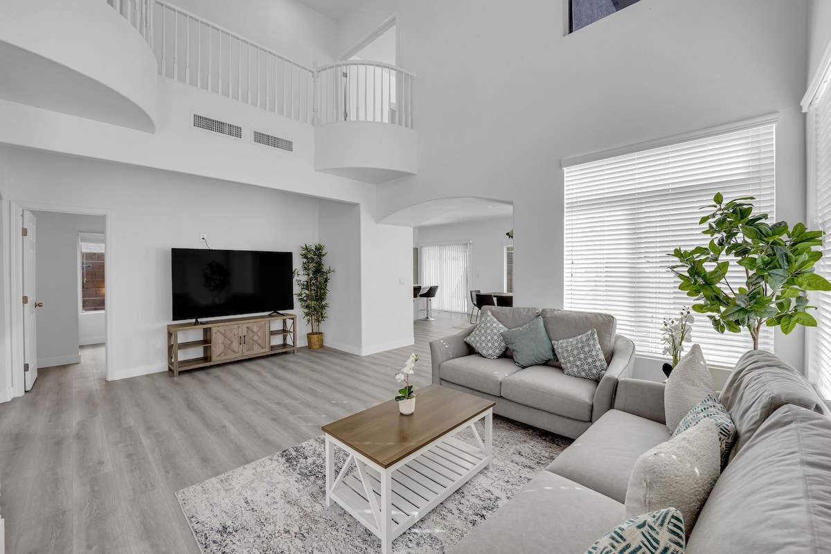 An open living space features a large light gray sectional sofa adorned with decorative pillows. A wooden coffee table rests on a soft area rug. A flat-screen TV is mounted on a light wooden media console. Natural light enters through large windows, complemented by greenery from potted plants.
