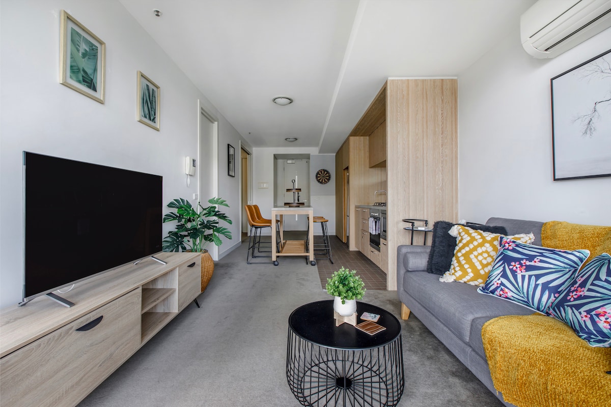 A modern living space features a plush sofa adorned with colorful cushions, a sleek coffee table, and a large television. Natural light fills the room through an adjacent area leading to the kitchen and dining space, where warm wooden furnishings create a welcoming environment.