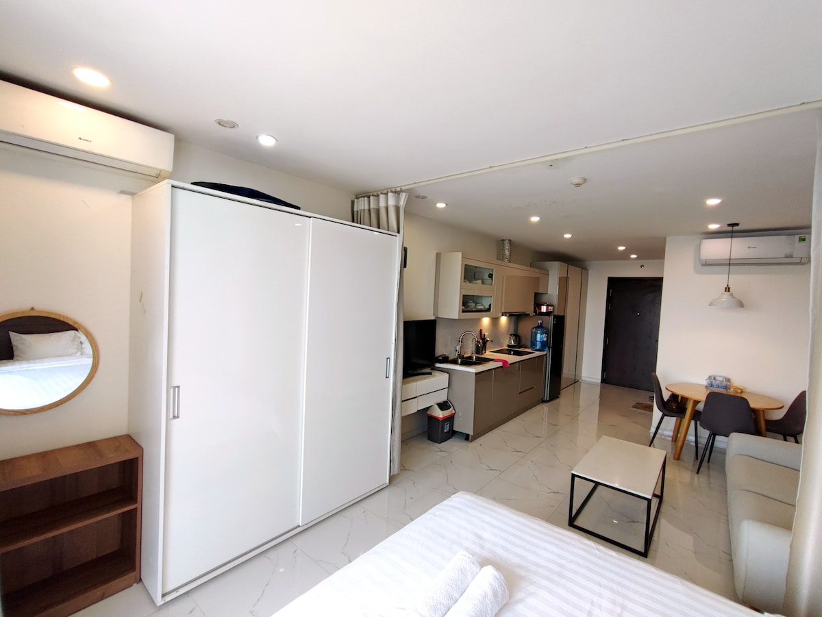 A well-lit studio apartment showcases an open layout featuring a comfortable sleeping area with a spacious bed. A compact kitchenette is visible, equipped with modern appliances, while a small dining area with a table and chairs is present. Storage options include a large wardrobe.