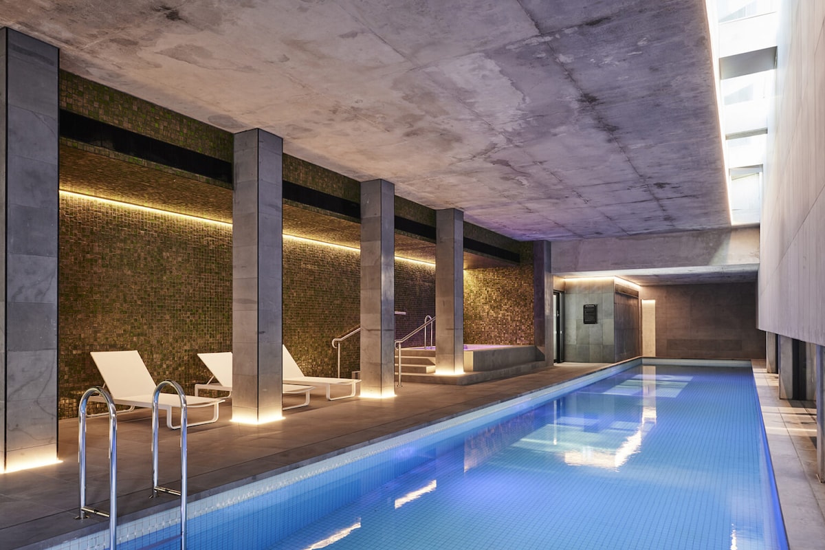 An indoor swimming pool area features a sleek, modern design with smooth concrete walls and green tiled accents. Two loungers are positioned alongside the pool, which is illuminated by soft lighting that reflects off the water's surface.