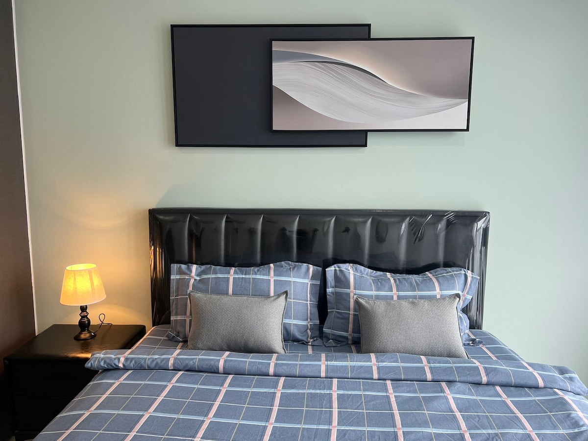 A comfortable sleeping area is highlighted by a spacious bed with a soft plaid blanket and decorative pillows. A bedside table holds a lamp, providing soft lighting. Above the bed, a modern artwork piece adds a touch of elegance to the serene wall.