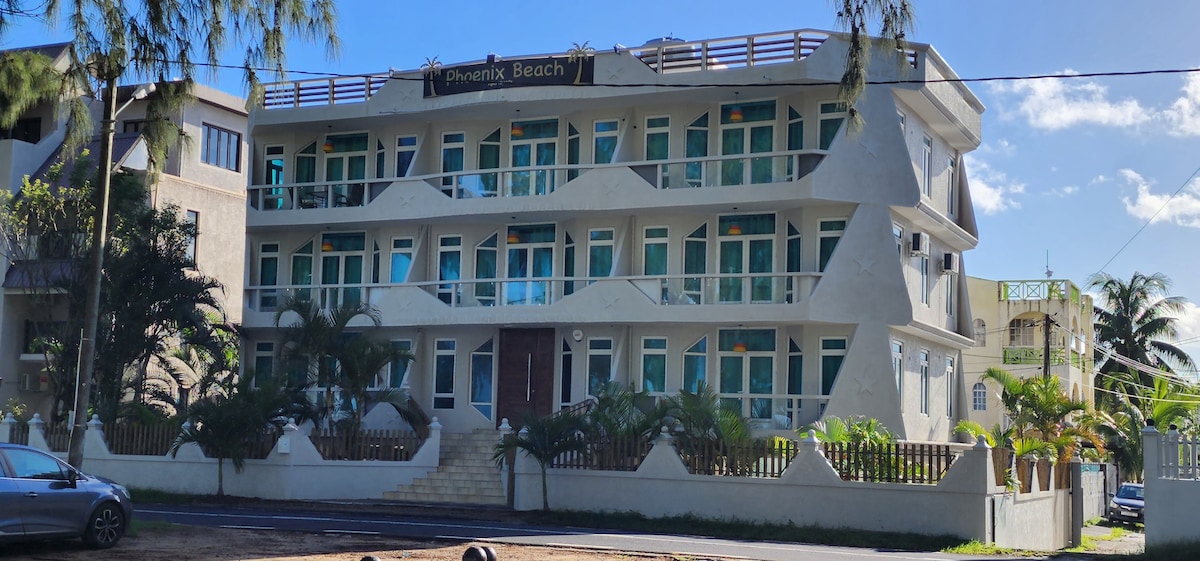 The exterior of a modern building is presented, featuring a light-colored facade with multiple large windows. Balconies are visible on the upper floors, adorned with railings. Lush greenery surrounds the entrance, contributing to a welcoming appearance along a tree-lined street.