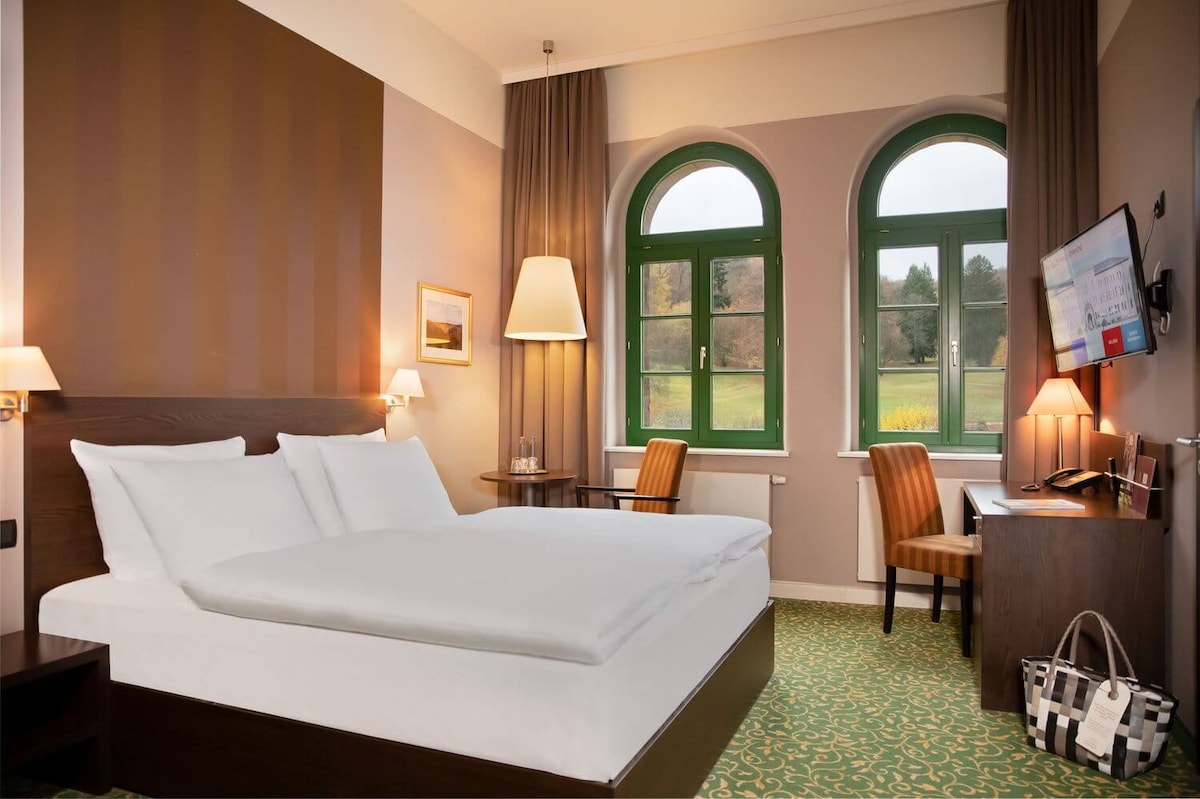 A spacious room is featured with a large, comfortable bed dressed in white linens. Two green arched windows allow natural light to illuminate the space. A desk and chair are positioned alongside a television. Earth tones and striped wallpaper create a calming atmosphere.