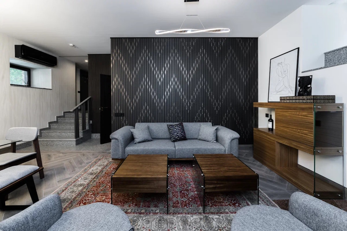 A modern living area features a gray sofa with accent cushions, two wooden coffee tables, and stylish armchairs. A patterned rug adds warmth, complemented by a sleek sideboard displaying decorative items. The wall is adorned with a geometric design, and a staircase leads to an upper level.