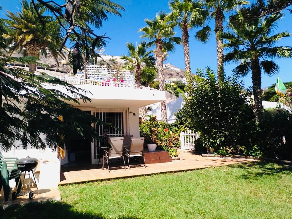 A serene outdoor space is surrounded by tropical palm trees and lush vegetation. Outdoor seating is positioned on a tiled patio, with a barbecue grill nearby. The grassy area adds to the inviting environment, offering a peaceful spot for relaxation.