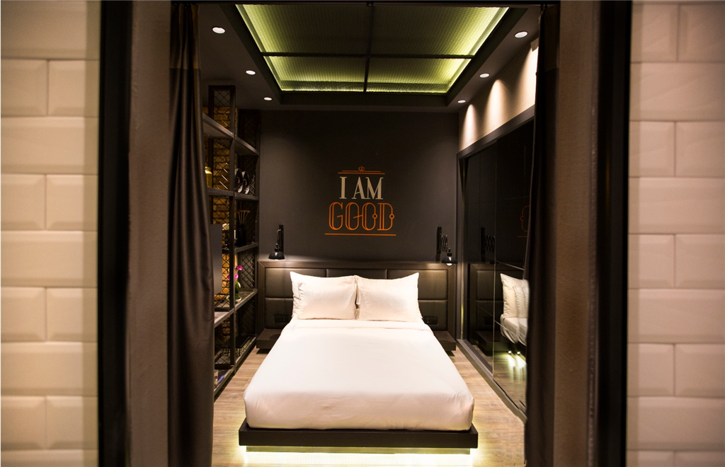 A modern bedroom features a spacious bed with crisp white linens. The room is accented by a dark wall displaying 'I AM GCGB' in bold letters. Mirrored surfaces and ambient lighting enhance the contemporary design, while shelves add storage and decorative elements.