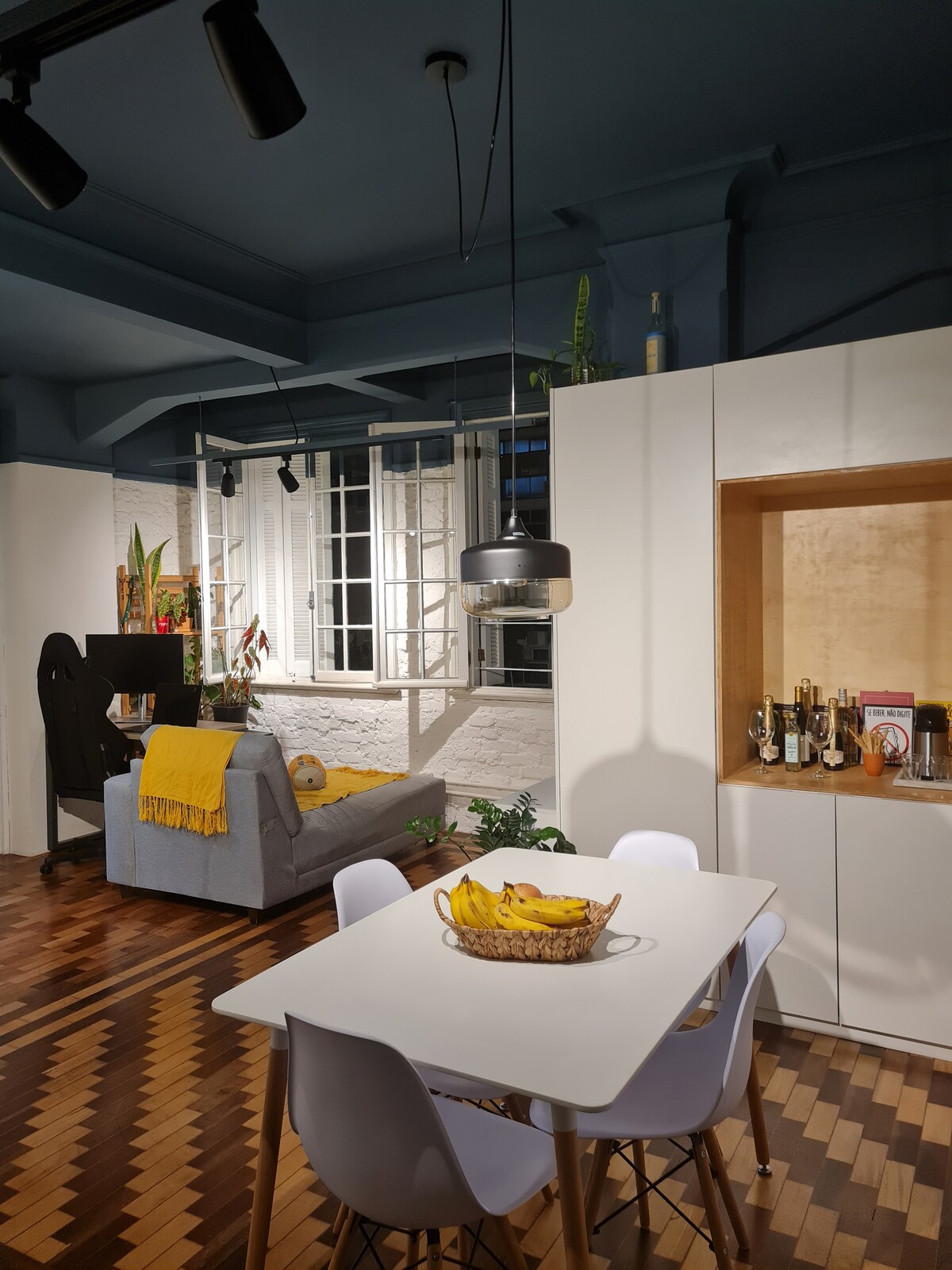 An open layout displays a dining area with a white table and four chairs, positioned near a large window. A cozy seating area features a gray sofa adorned with a yellow throw. Plants are placed around the space, enhancing the inviting environment.