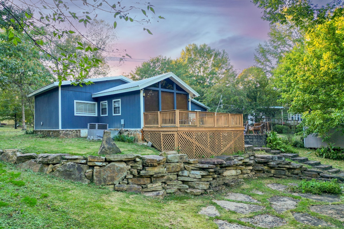 The cabin's exterior showcases a blue façade complemented by a wooden deck. A stone pathway leads through a lush green area with trees providing natural shade. The peaceful setting features a gentle slope and inviting outdoor space for relaxation.