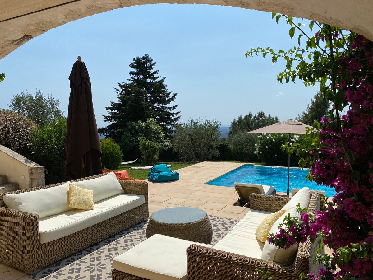 A shaded terrace features comfortable seating arrangements, accompanied by blooming flowers. In the background, a private pool is visible, surrounded by lush greenery and an olive tree. The area offers expansive views of the scenic landscape.