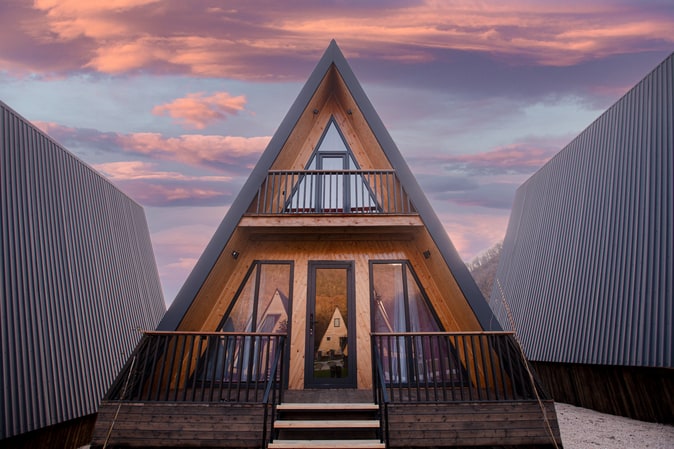 A distinctive triangular-shaped building features a wooden exterior and large front windows. A balcony extends above the entrance, providing views of the surroundings. The colorful sunset sky creates a dramatic backdrop for this unique architectural design.