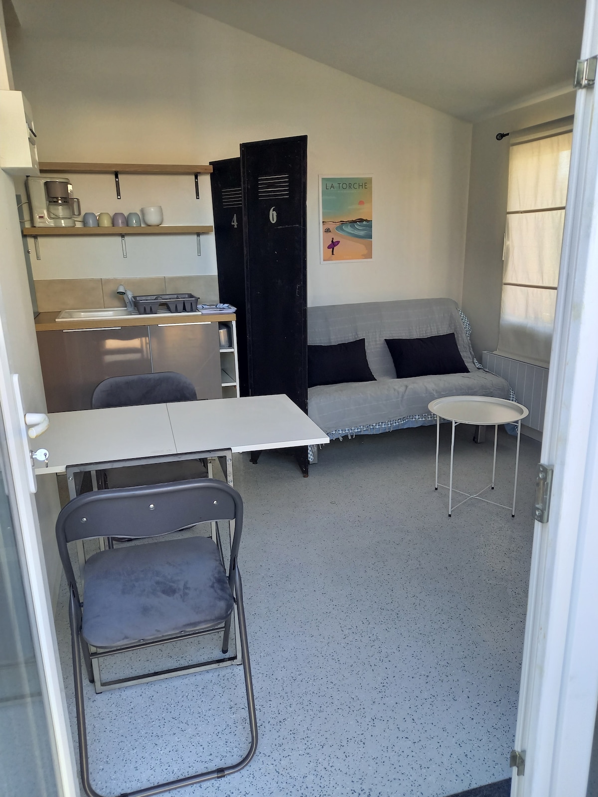 A bright living area features a gray sofa positioned beside a small round table with a chair. In the background, a kitchen counter is equipped with dishes and appliances, while a piece of wall art adds a touch of color. Natural light streams through a partially open window.