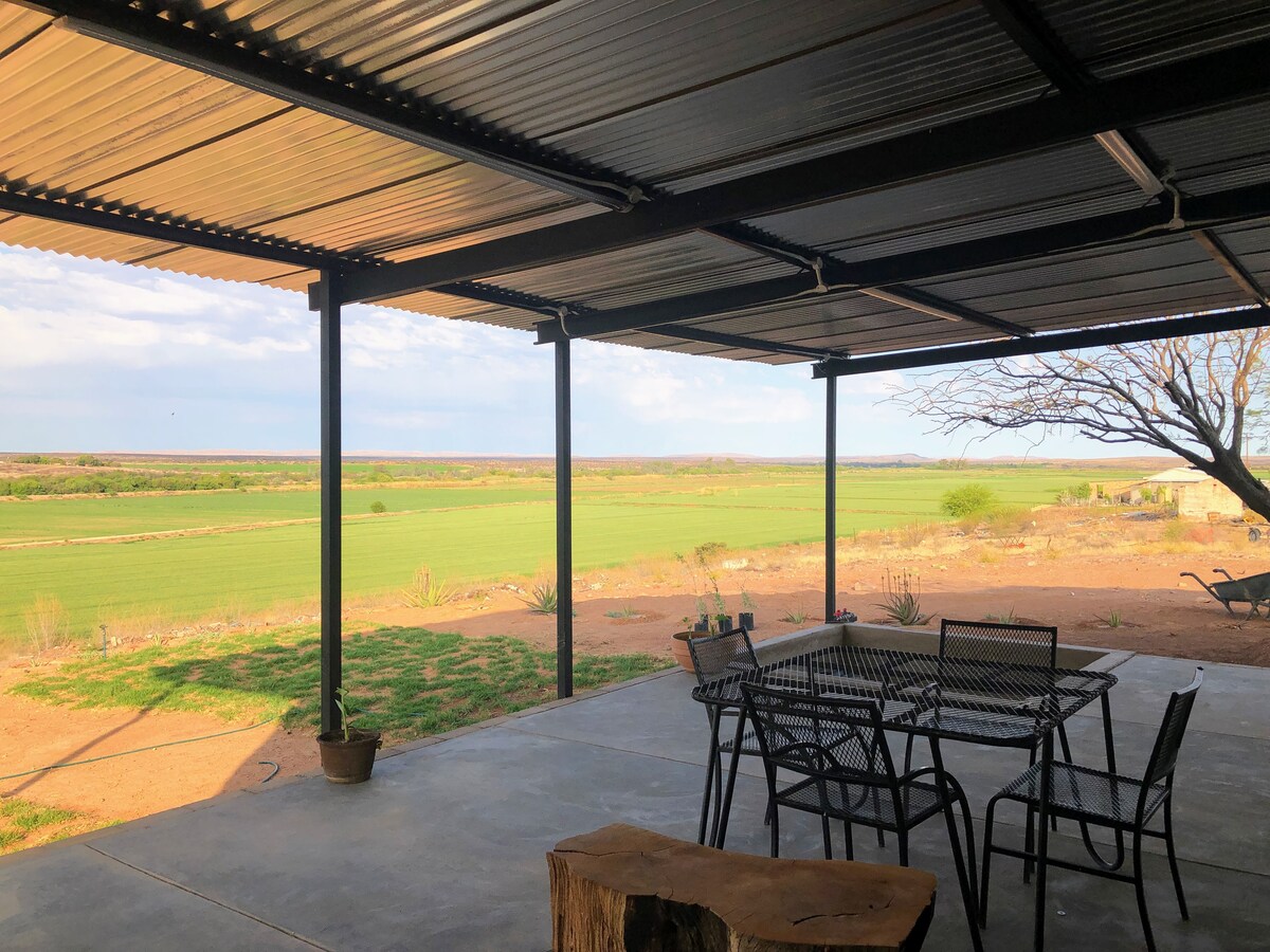An outdoor seating area features a metal table surrounded by several black chairs, positioned under a corrugated roof. Expansive green fields stretch across the horizon, framed by a clear sky, with gentle shadows cast on the concrete floor.