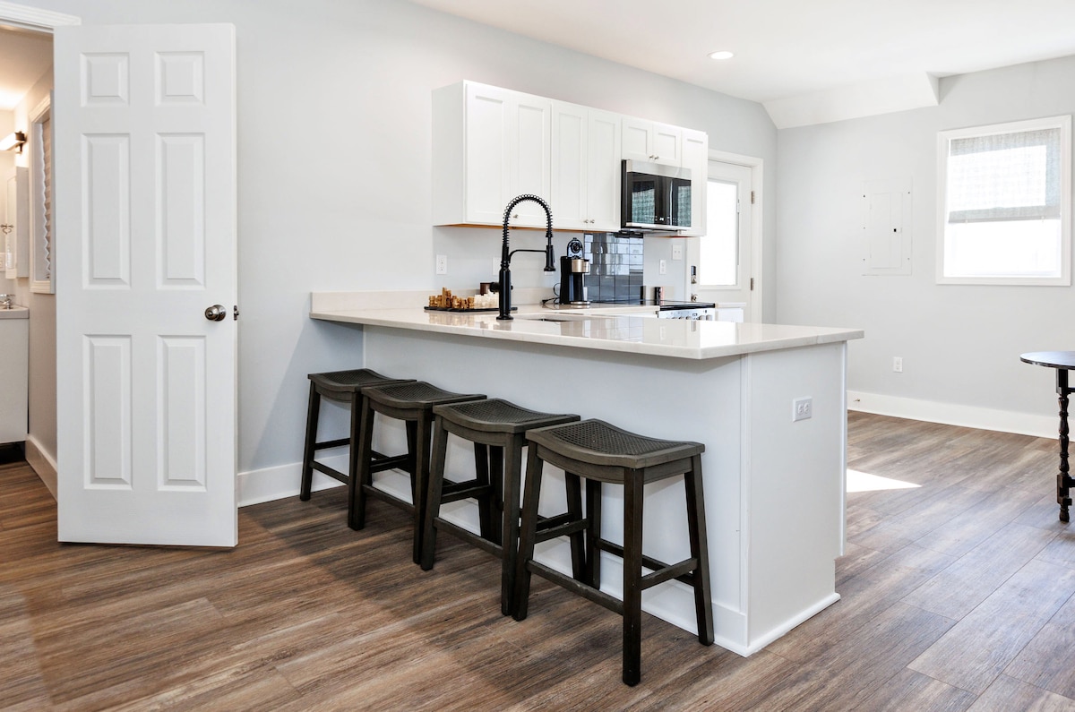A modern kitchen area is highlighted by a spacious countertop and a set of four dark wooden stools. White cabinetry and stainless-steel appliances offer a clean aesthetic, while natural light floods in through a large window, illuminating the light grey walls and hardwood flooring.