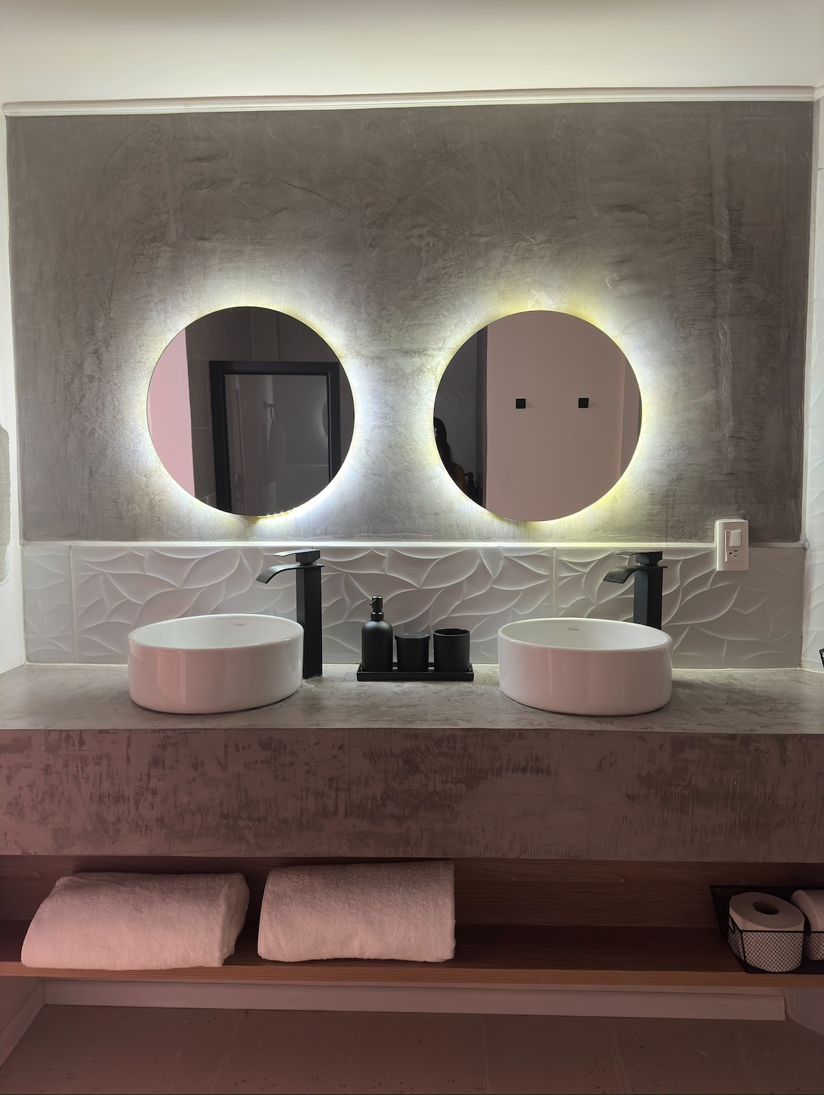 A modern bathroom features two round mirrors with soft backlighting above a countertop shared by two sleek, white vessel sinks. Below, neatly arranged towels are placed on a wooden shelf, complementing the elegant design elements of the space.