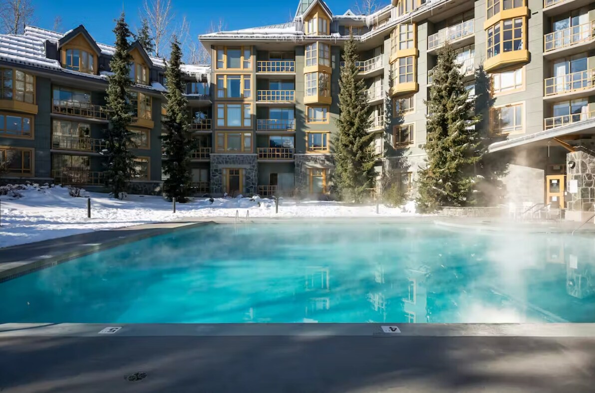 A heated outdoor pool is featured in the foreground, surrounded by snow and towering evergreen trees. The modern lodge architecture is visible in the background, showcasing large windows and a welcoming entrance. Steam rises gently from the pool, set against a clear blue sky.