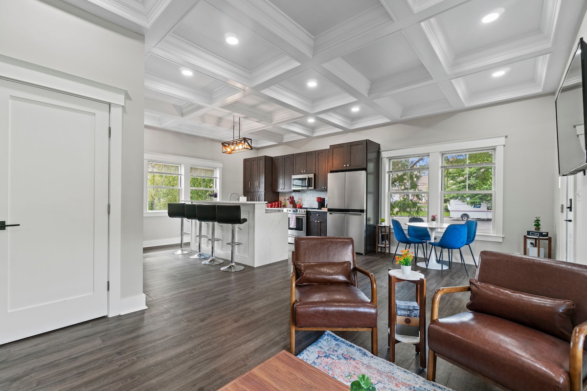 A spacious open-concept living area features a modern kitchen with stainless steel appliances and a central island. Comfortable seating options include two armchairs and a sofa, complemented by a dining table and blue chairs. Natural light pours in through large windows, enhancing the inviting space.