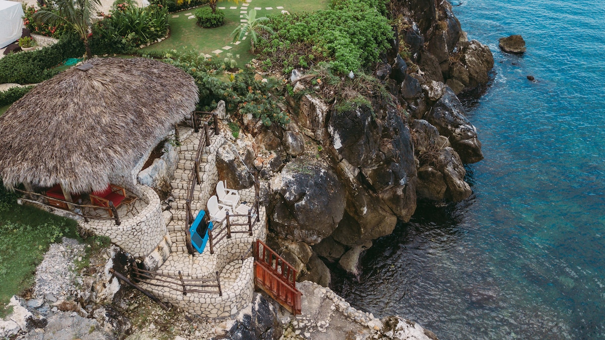 An aerial view reveals a rustic cabana with a thatched roof, surrounded by lush greenery. Stone pathways lead to the water's edge, where a rocky shoreline meets clear waters. Lounge chairs are placed near the cabana, offering a serene spot to enjoy the ocean views.