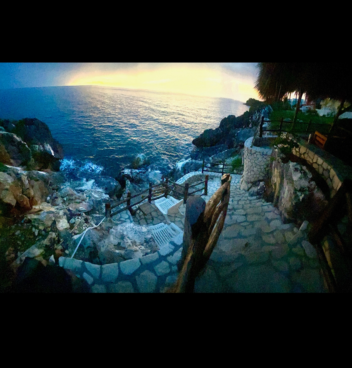 A stone pathway leads down to the rocky shoreline, bordered by wooden railings. Waves gently crash against the shore under a soft twilight sky, while the tranquil sea reflects shades of blue and purple as the day transitions into evening.