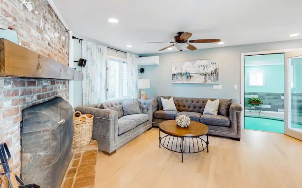 A well-lit living room features a comfortable gray sectional sofa with tufted detailing, arranged around a round coffee table. A rustic brick fireplace is visible, complemented by a large window that brings in natural light, leading to the indoor pool area.