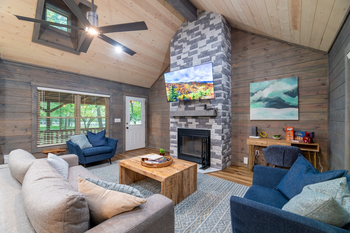 The main living area features a stone fireplace as a focal point, with a mounted TV above. Two blue armchairs and a comfortable gray sofa surround a wooden coffee table, all set against a backdrop of wooden walls and a high, sloped ceiling. Natural light filters in through large windows.