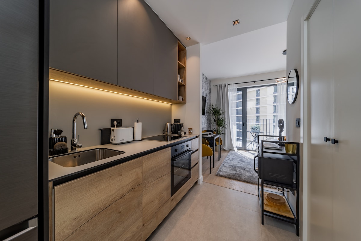 A modern kitchen features sleek cabinetry with a mix of dark wood and light finishes. Stainless steel appliances are positioned conveniently, with a double-basin sink and ample counter space. Natural light enters through large windows, illuminating a cozy dining area with stylish chairs.