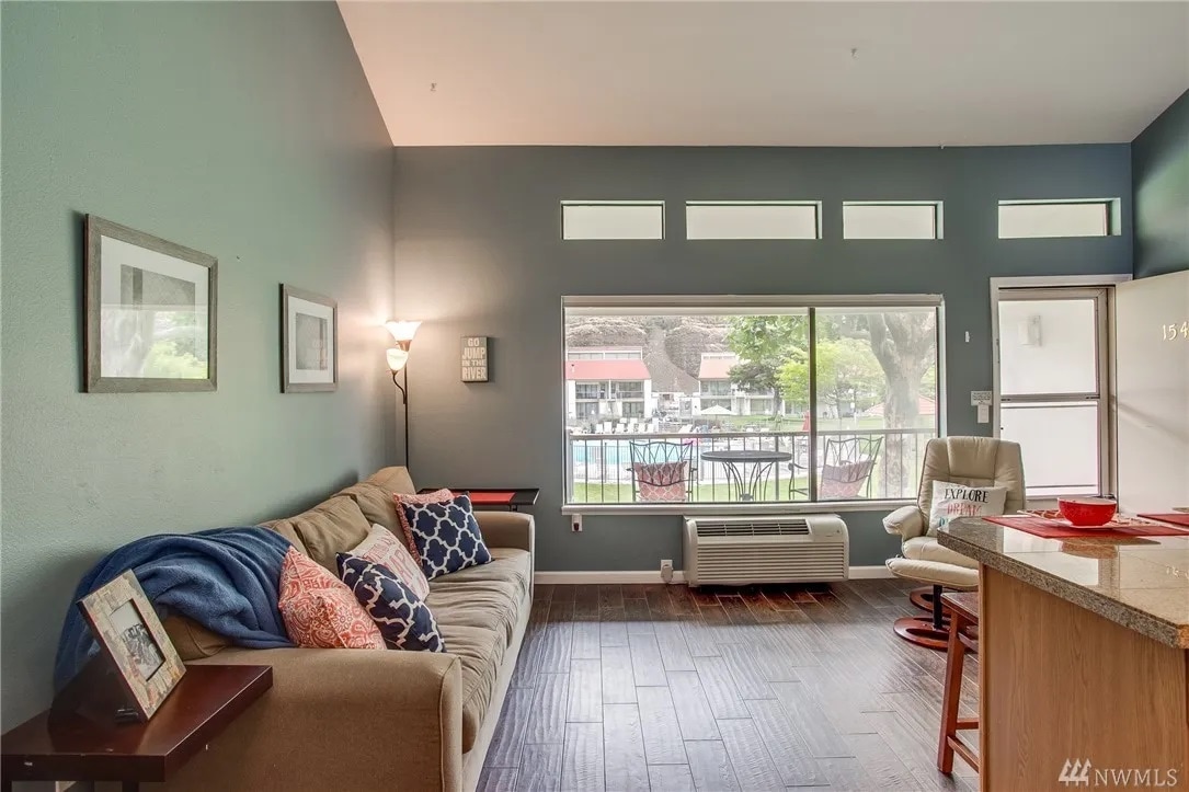 A living area features a comfortable beige sofa with a blue throw blanket, positioned near large windows that provide natural light. Decorative frames are displayed on the gray walls. A small kitchen island with seating is visible, offering a welcoming space.