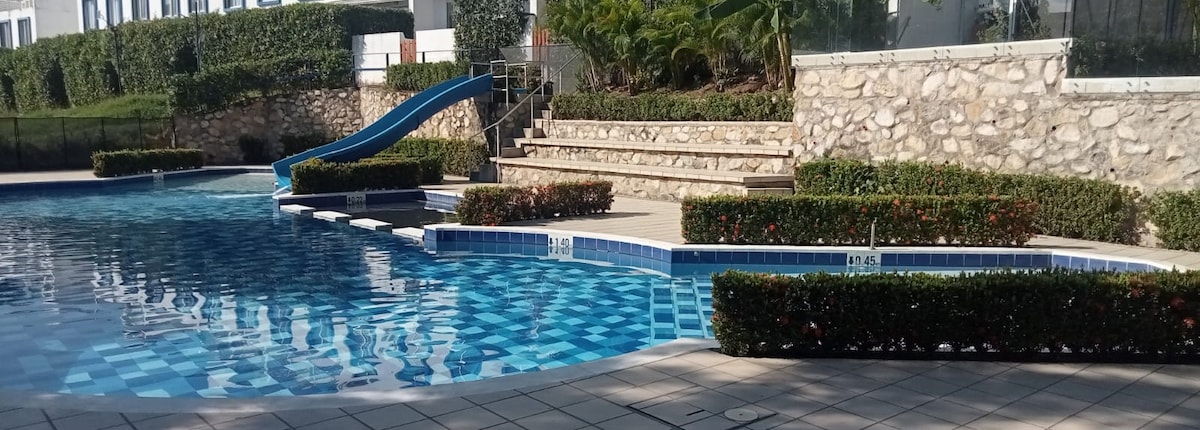 A spacious pool area features a blue water slide leading into the inviting pool. Surrounding the pool are well-maintained gardens with hedges and flower beds. Stone steps provide access to the elevated seating area, offering a relaxing environment for guests.
