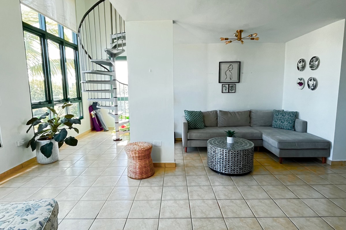 A bright living area is highlighted by large windows allowing natural light to fill the space. A spiral staircase leads to an upper level. The corner seating arrangement features a gray sofa with decorative pillows, complemented by a patterned coffee table and a small woven stool.