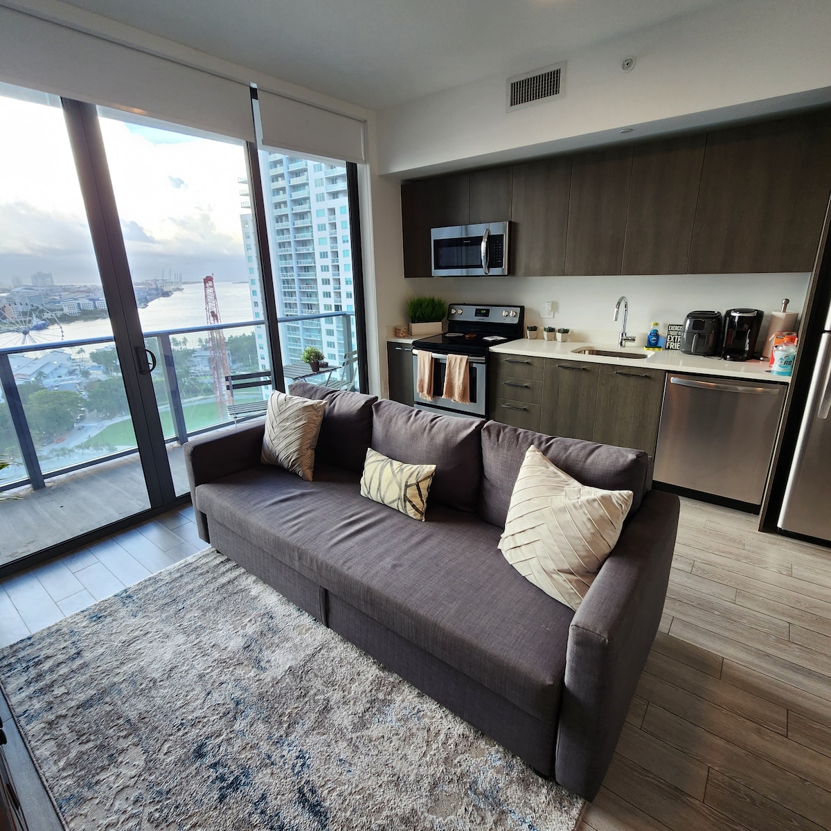 A modern studio apartment is showcased, featuring a spacious sofa with decorative pillows facing a glass balcony overlooking the water. The kitchen area is visible, equipped with stainless steel appliances, cabinetry, and countertop space, enhancing functionality and style.