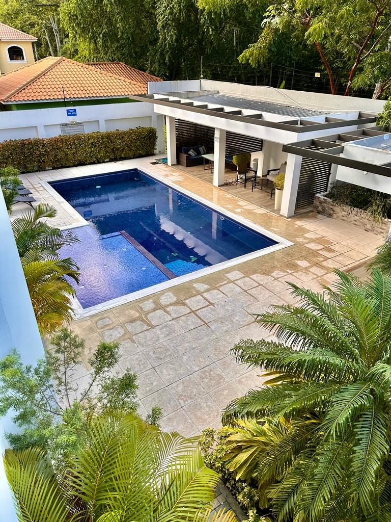 A shared outdoor pool is surrounded by lush greenery and well-maintained tile flooring. A shaded seating area with a table and chairs is positioned near the pool, providing a comfortable space for relaxation. The area is enclosed by a low wall and features vibrant tropical plants.