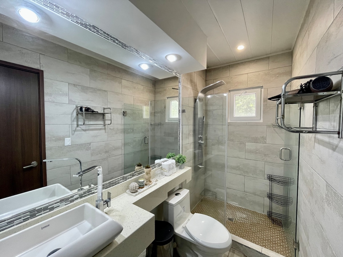 The bathroom is equipped with modern fixtures and a spacious shower area enclosed in glass. A double sink with a minimalist countertop is featured, complemented by contemporary shelving and subtle greenery. Natural light enters through a small window, enhancing the overall brightness of the space.