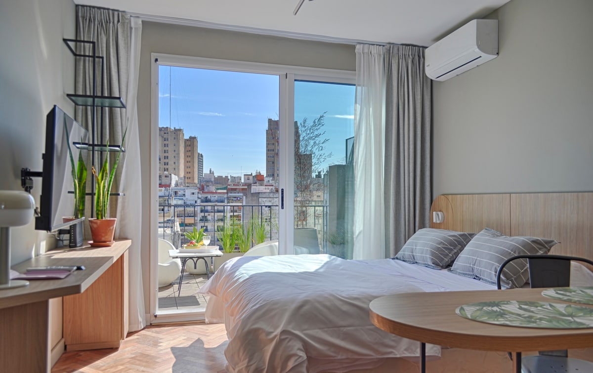 A bright studio is shown with large glass doors leading to a private terrace. A spacious bed is positioned against a wooden headboard, and a small dining area is featured with a round table. Plant life is visible outside, complementing the urban view.