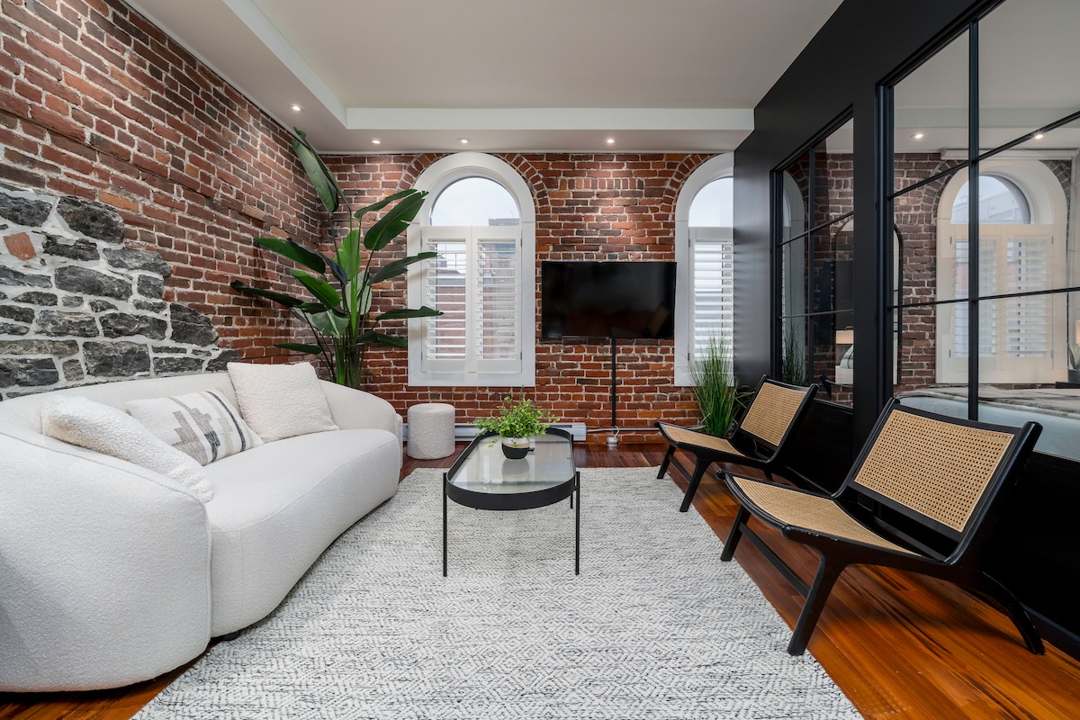 A spacious living area features striking red brick walls paired with a contemporary design. A comfortable white sofa is positioned around a modern black coffee table, with two stylish chairs nearby. Plants add a touch of greenery, and windows allow natural light to fill the space.