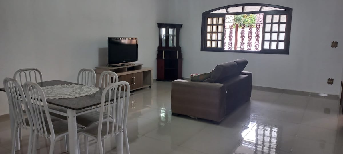 A spacious living area features a comfortable sofa and a dining table with six chairs. A television is positioned on a cabinet, and a window with decorative grills allows natural light to fill the room. The glossy floor reflects the soft illumination of the space.