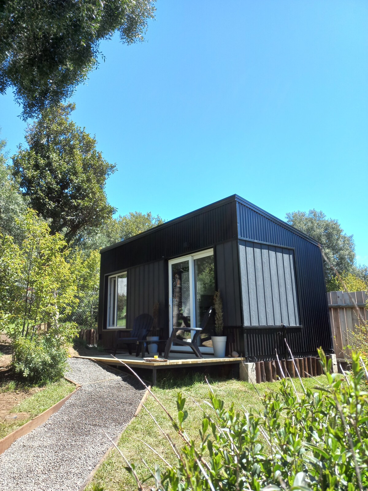 A contemporary tiny house features a dark exterior with large windows that allow natural light to fill the interior. A small porch is visible, furnished with two chairs and a potted plant. Lush greenery surrounds the structure, enhancing the peaceful outdoor environment.
