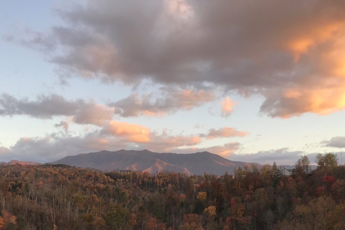 A sweeping view of the Great Smoky Mountains is captured at sunset, showcasing a rich tapestry of autumn foliage in shades of orange, yellow, and red. Wispy clouds drift across the sky, hinting at the tranquil beauty of the natural landscape below.