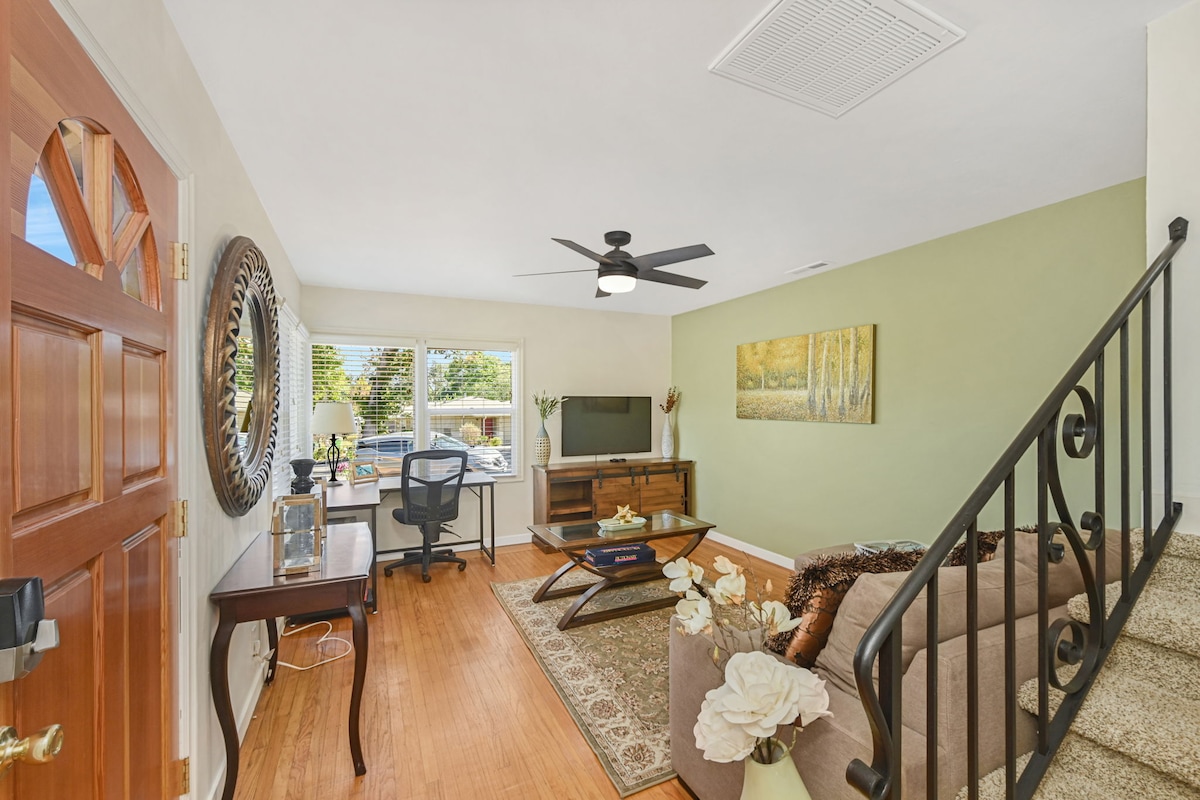 The living area features a spacious sectional sofa paired with an area rug, complemented by a coffee table. A flat-screen TV is mounted on the wall, and a work desk is positioned near the window, providing natural light. Stairs lead to the upper level.