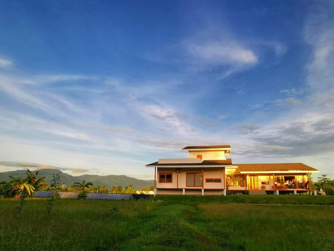 A modern house is positioned amidst lush greenery, featuring multiple windows that reflect natural light. The surrounding landscape includes rolling hills in the background and a clear blue sky. A path of grass leads toward the entrance, emphasizing the serene and expansive setting.