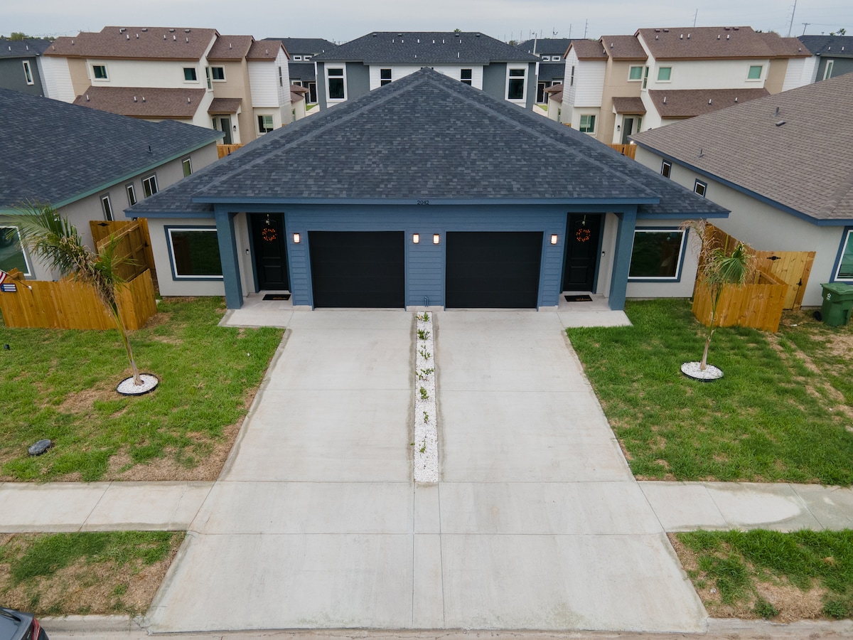 An aerial view showcases a modern duplex with a symmetrical façade, featuring two separate entrances. The driveway leads to black garage doors, framed by decorative palm trees and well-maintained landscaping. A central pathway draws attention to the entrance, enhancing the property’s welcoming appeal.